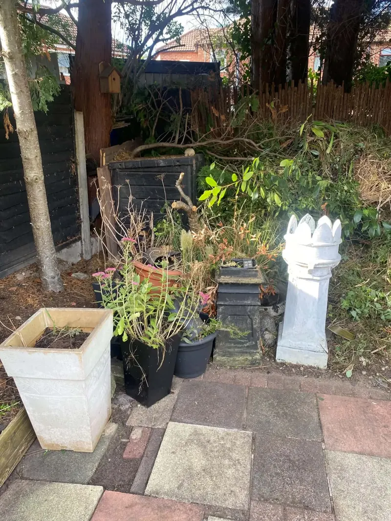 White chimney-pot ornament, planters and cut branches in an untidy corner of a garden beside a dark fence