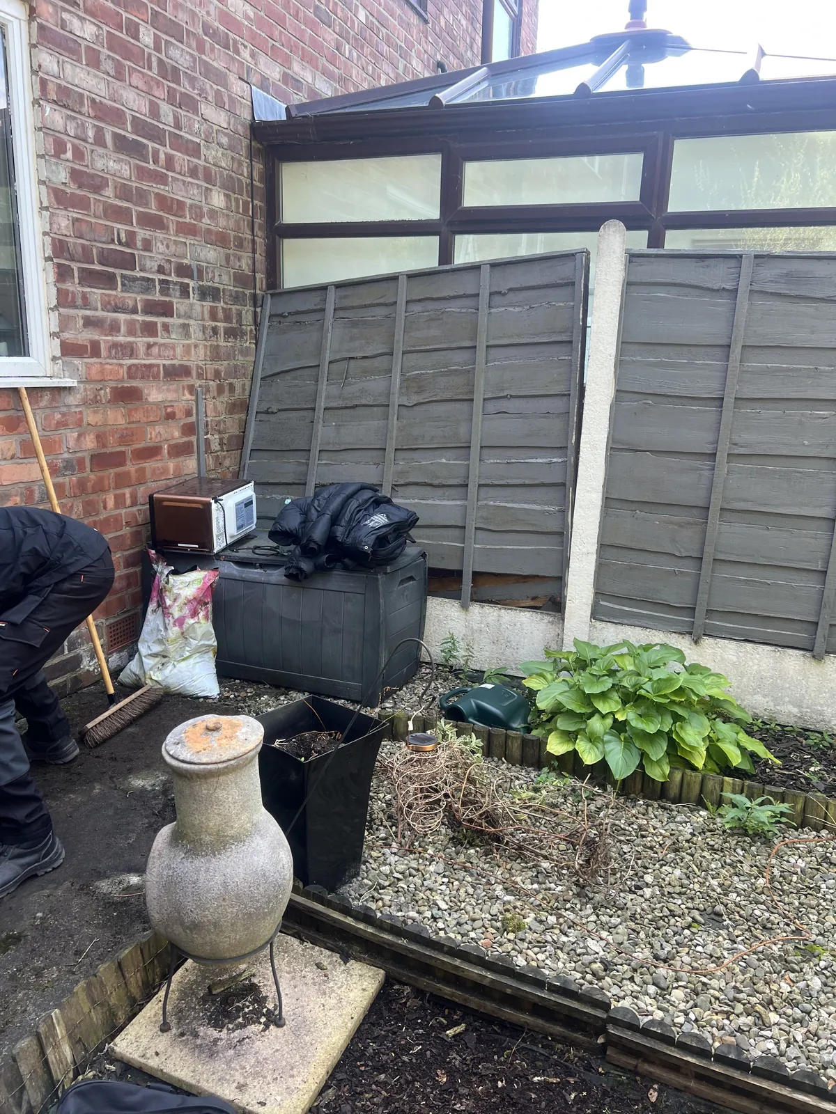 Side of a brick house with a conservatory, a leaning grey fence panel, a storage box and bags against the wall, a gravel bed with a hosta and a person in dark work clothes leaning over at the left edge