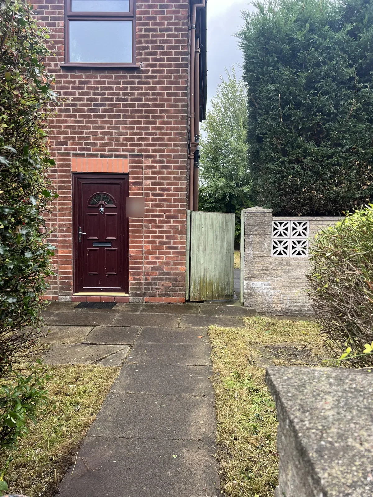 Front path of a brick house with a dark red door, paving slabs, short cut lawn either side and trimmed shrubs framing the path, the house number hidden