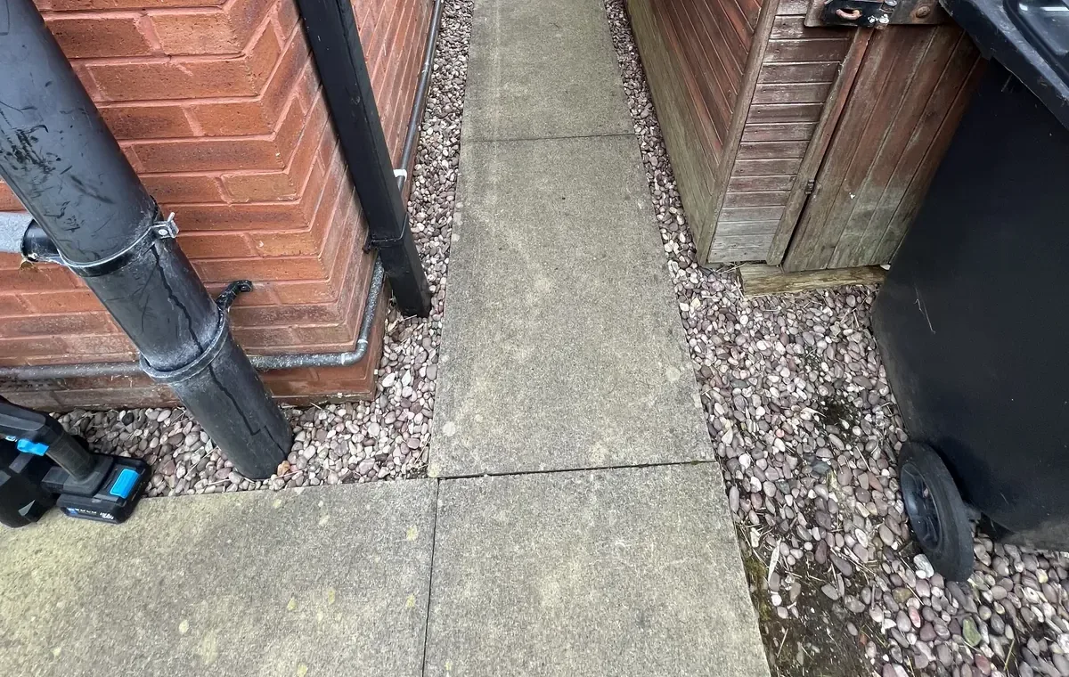 Downpipe running into the ground beside a slab path and gravel strip, with a shed and wheelie bin opposite