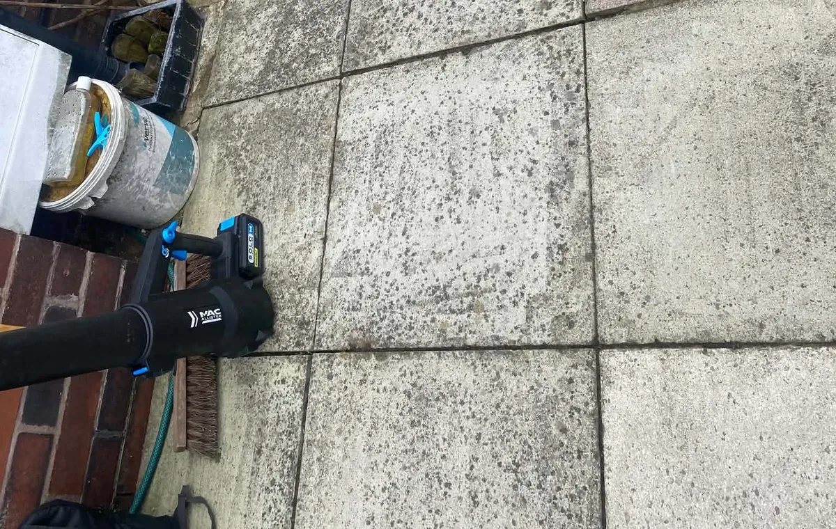 Battery leaf blower and a stiff broom on a grey slab patio beside planters and a raised bed