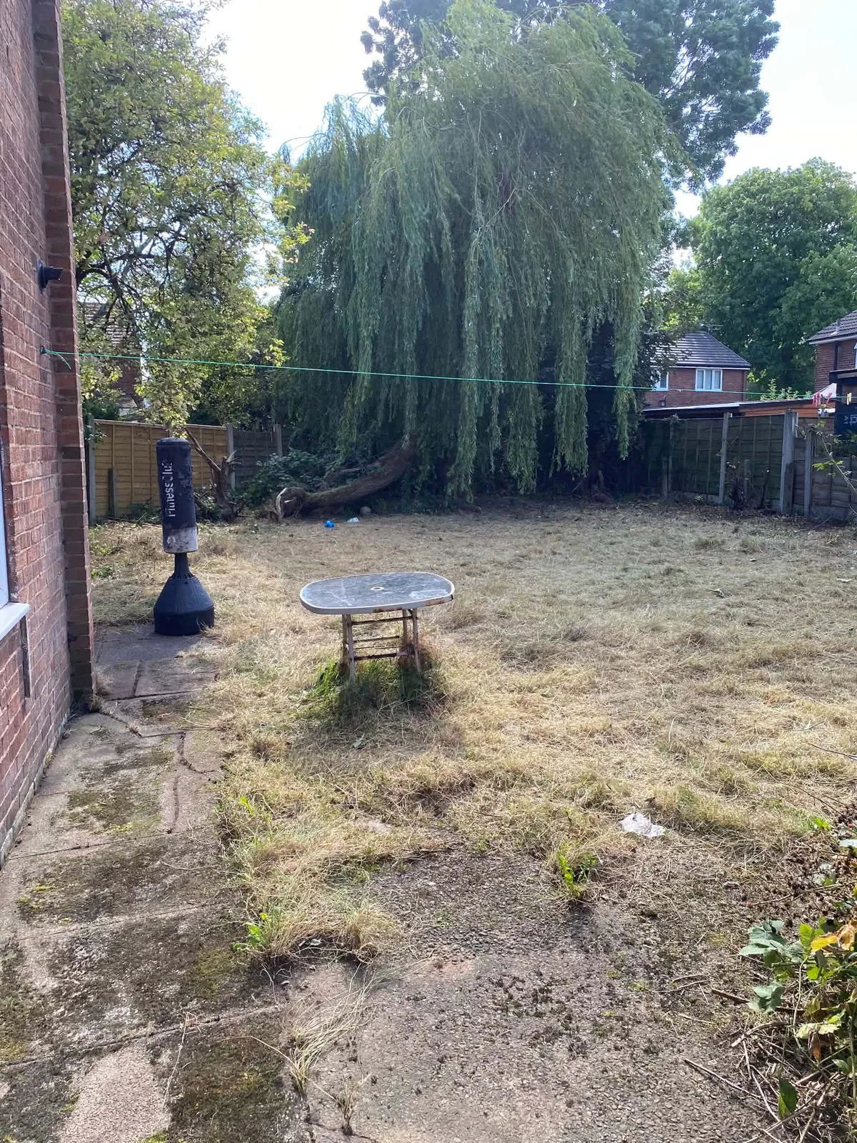 Old paving slabs along a brick house wall at the edge of a cut lawn, with a metal table and a willow tree behind