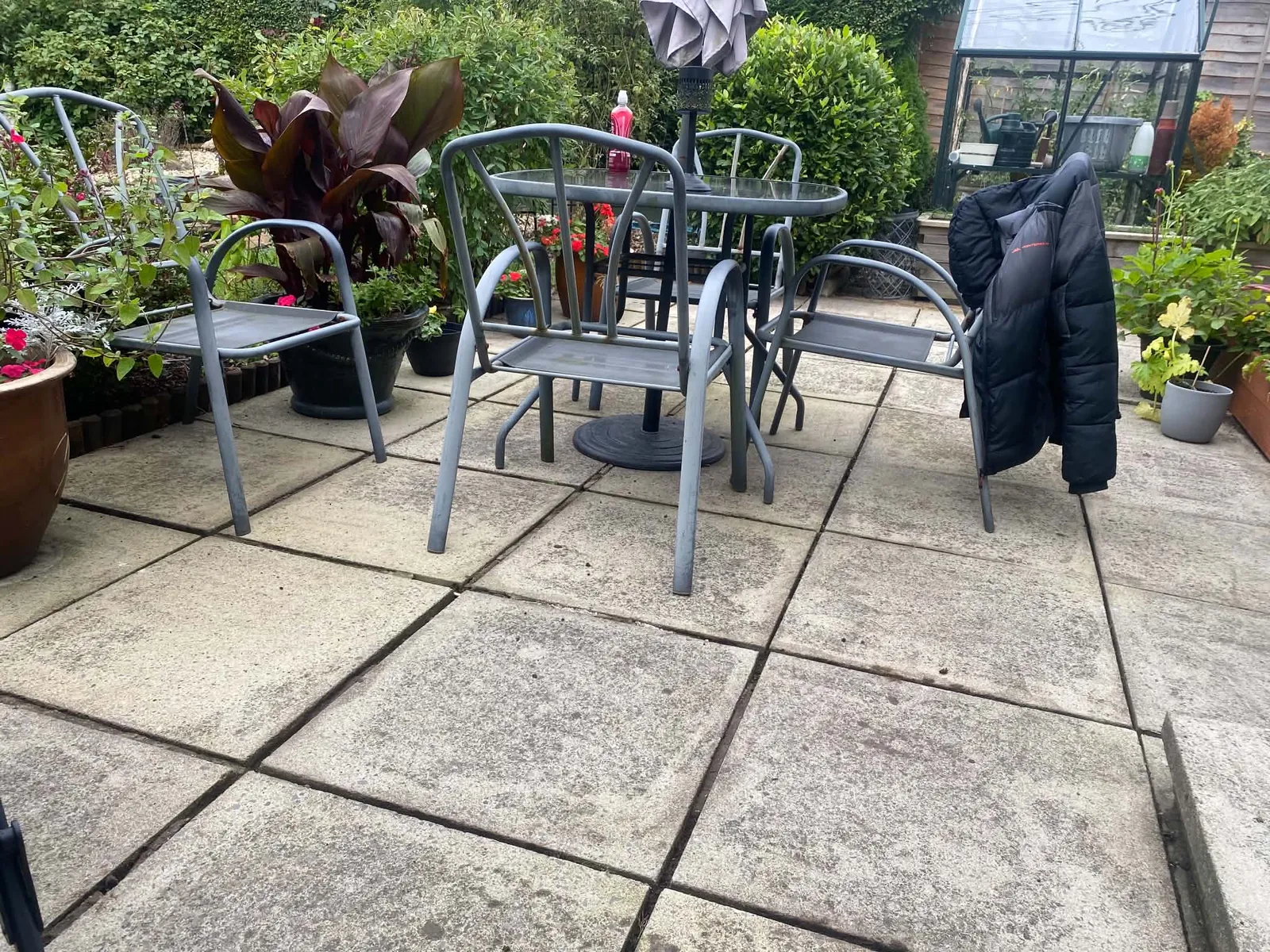 Pale grey paving slabs on a small garden patio with a metal table and chairs, planters and a greenhouse behind, the slabs clean with light mottling