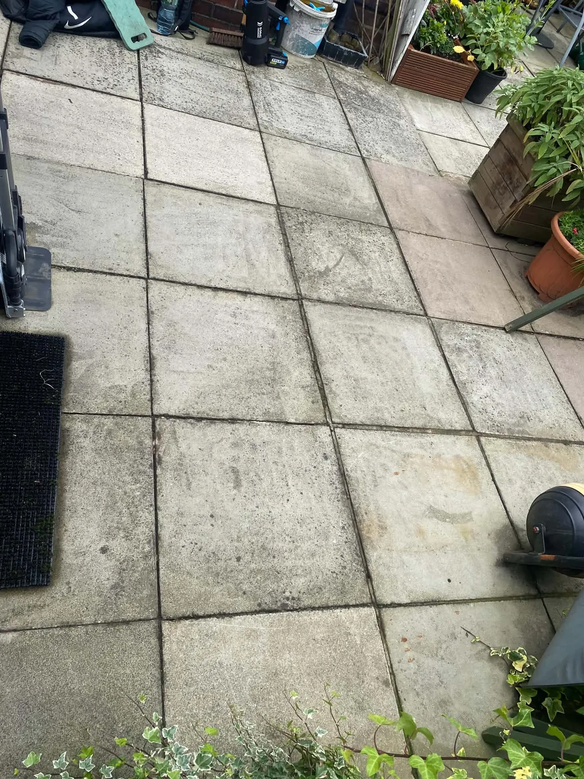 Grey concrete paving slabs on a patio with dark staining in the joints and on some slabs, a pressure washer at the left edge and planters at the top right
