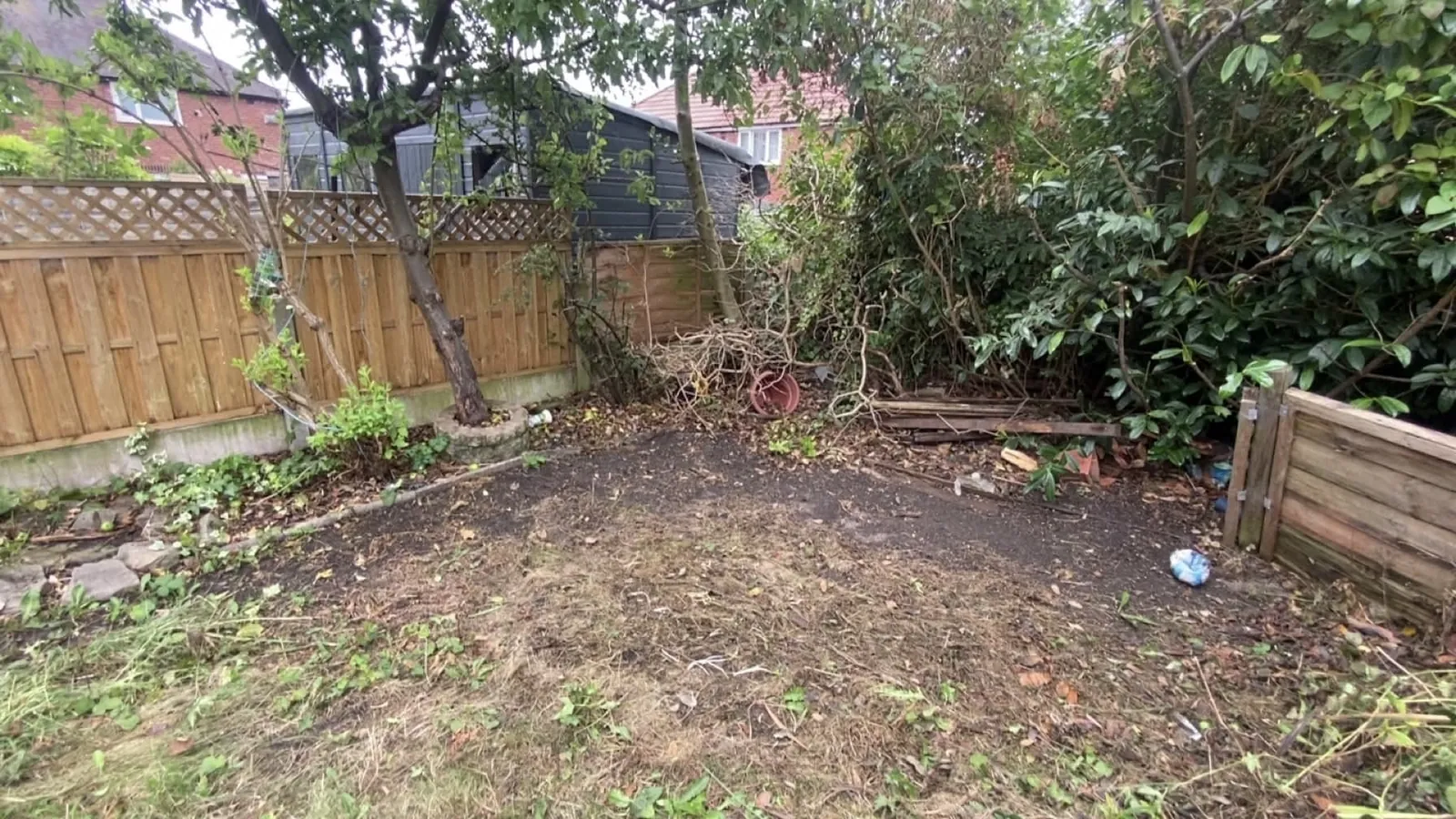 Cleared back garden with bare ground, a timber fence and thick shrubs at the edge