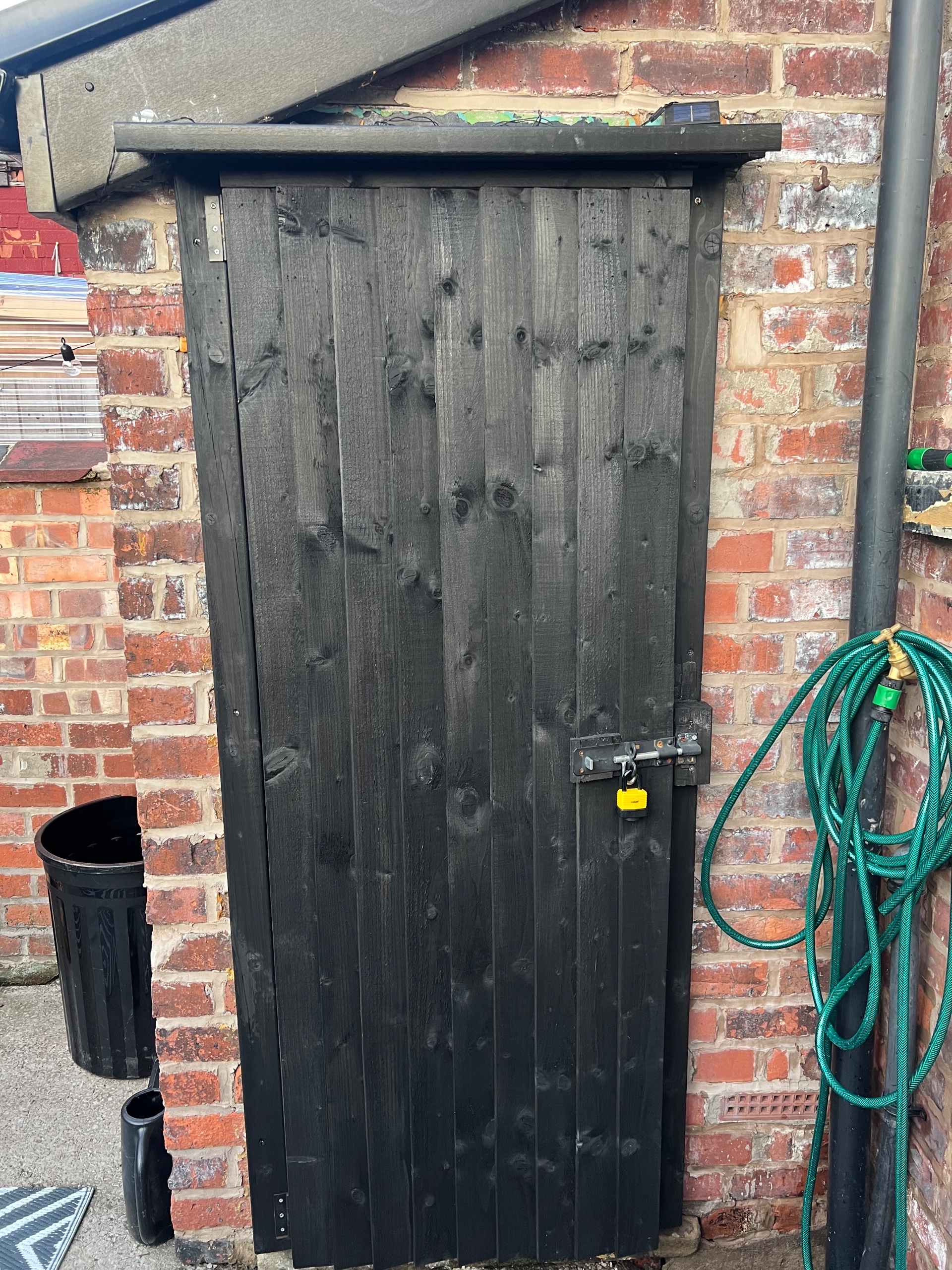 painted black timber garden cupboard door with padlock against brick wall and garden hose
